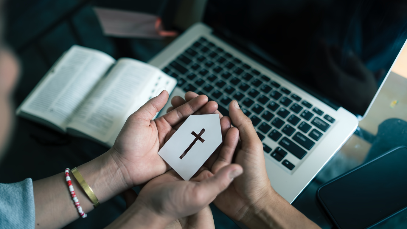 Hands holding a cross-shaped card, Bible and laptop in the background.